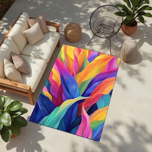 May include: A rectangular outdoor rug with a vivid, abstract design of colourful leaves in shades of blue, purple, yellow, orange, and pink. The rug is placed on a light-coloured patio, near a white sofa and potted plants.