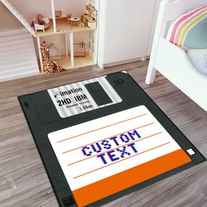 May include: A black rectangular rug designed to resemble a 3.5-inch floppy disk. The rug features the text "CUSTOM TEXT" in a pixelated font, along with the "imation 2HD IBM" logo and specifications. The bottom edge has an orange stripe.