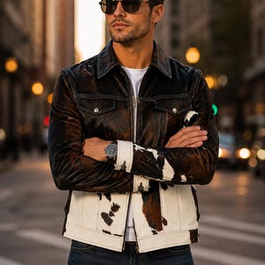 May include: A jacket featuring a dark brown and black textured upper section and a white, brown, and black cowhide print lower section. The garment has a pointed collar, a front zipper closure, two chest pockets with button flaps, and long sleeves.
