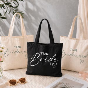 Puede incluir: Dos bolsas de tela color hueso con el texto Team Bridesmaid y un pequeño corazón, y una bolsa de tela negra con el texto Team Bride y un corazón. Cada bolsa cuenta con dos asas para el hombro y una forma rectangular.