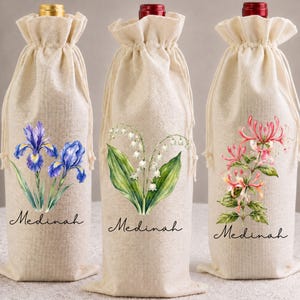 May include: Three beige linen wine bottle bags with drawstring closures. Each bag features a botanical illustration: a blue iris, white lily of the valley, and pink honeysuckle. The name Medinah is printed in black script below each floral design.