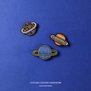May include: Three embroidered planet patches featuring a dark blue planet with gold swirls, a rainbow-striped planet, and a solid blue planet with a green ring. The text STITCHED CENTURY HANDWORK and HAND EMBROIDERED is printed at the bottom.