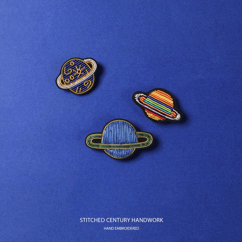 May include: Three embroidered planet patches featuring a dark blue planet with gold swirls, a rainbow-striped planet, and a solid blue planet with a green ring. The text STITCHED CENTURY HANDWORK and HAND EMBROIDERED is printed at the bottom.
