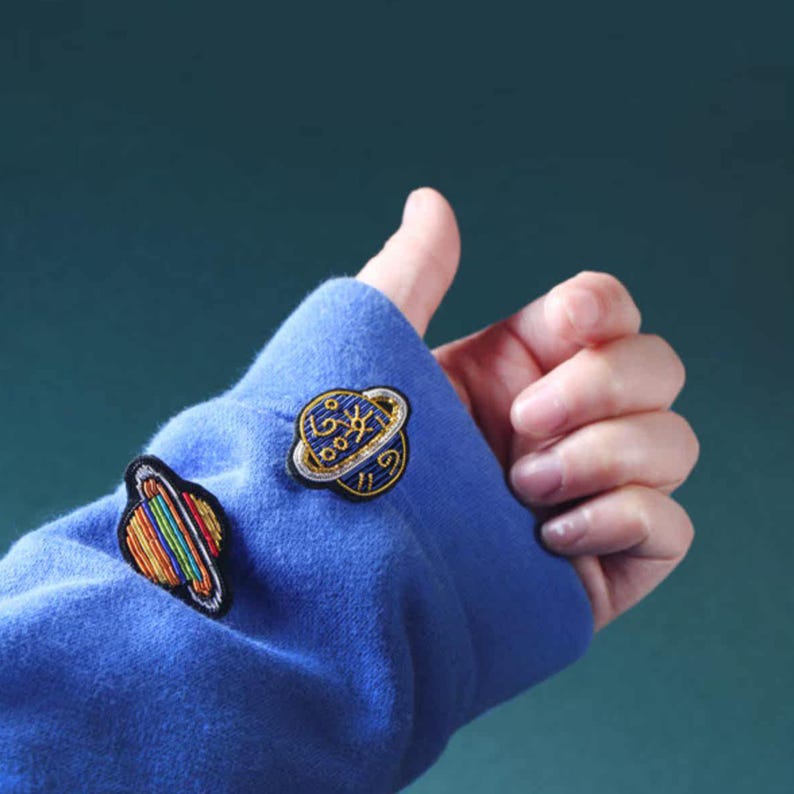 May include: A blue long-sleeved garment featuring two embroidered planet patches. One patch is a dark blue sphere with gold celestial symbols and a ring. The second patch is a multi-colored striped sphere with a ring, showcasing a rainbow pattern.