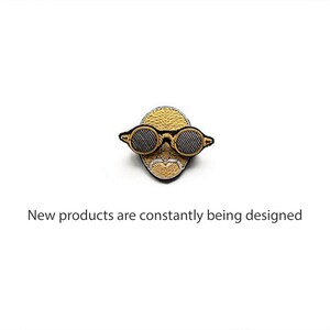 May include: An embroidered patch featuring a stylized golden face wearing round black-rimmed glasses, accompanied by the text "New products are constantly being designed" positioned directly below the embroidered figure on a plain white background.