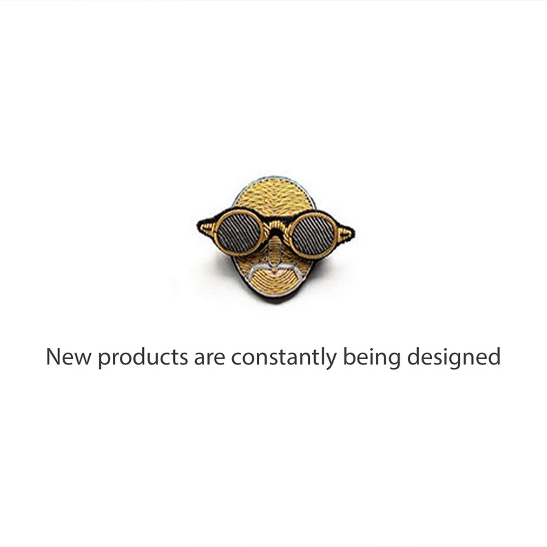 May include: An embroidered patch featuring a stylized golden face wearing round black-rimmed glasses, accompanied by the text "New products are constantly being designed" positioned directly below the embroidered figure on a plain white background.