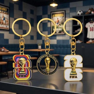 May include: Three FIFA World Cup 2022 keychains. One keychain has a colorful design, another has a black and gold circular design, and the third has a white and gold design. Each keychain features a gold-colored ring and the FIFA logo.