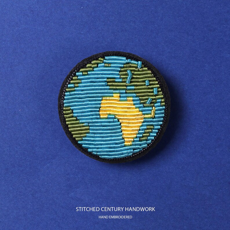 May include: A circular embroidered patch featuring a stylized globe design. The continents are rendered in yellow and green thread, while the oceans are depicted in horizontal rows of light blue thread. The patch has a black border and text below.