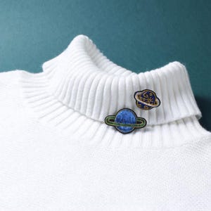 May include: A white ribbed turtleneck sweater featuring two embroidered planet-shaped patches. One patch is a blue planet with a green ring, and the other is a dark blue planet with a yellow ring and small yellow celestial symbols.