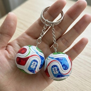 May include: Two miniature soccer ball keychains. Each ball features a white base with red, blue, and green curved designs, and a blue star pattern. The keychains have silver metal rings and chains.