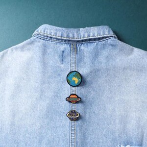 May include: Three circular embroidered patches featuring space themes attached to the back of a light blue denim jacket. The top patch depicts the Earth, the middle patch shows a ringed planet with orange and blue stripes, and the bottom patch is a blue planet.