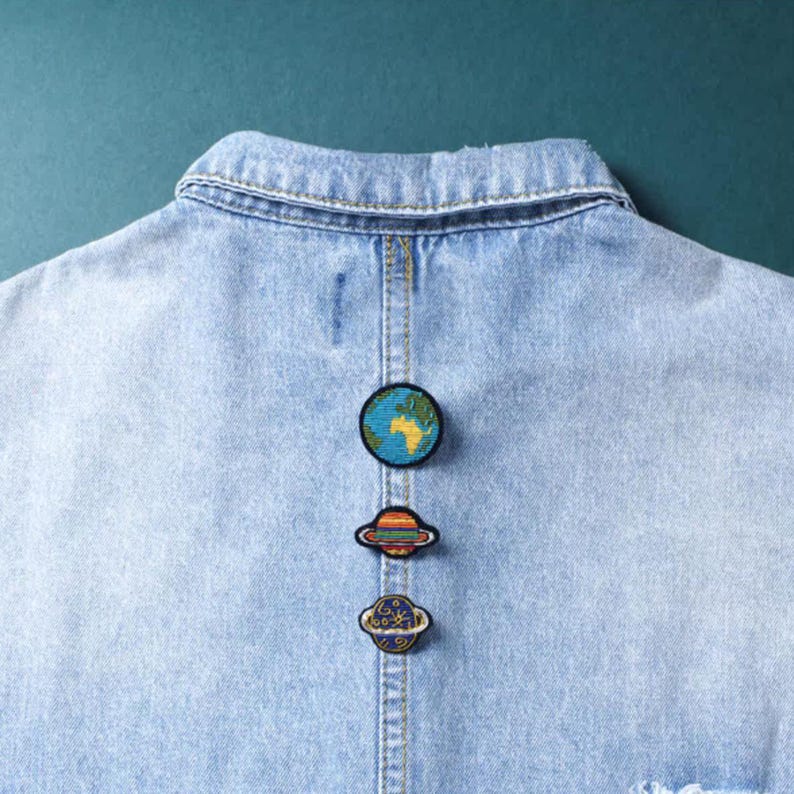 May include: Three circular embroidered patches featuring space themes attached to the back of a light blue denim jacket. The top patch depicts the Earth, the middle patch shows a ringed planet with orange and blue stripes, and the bottom patch is a blue planet.