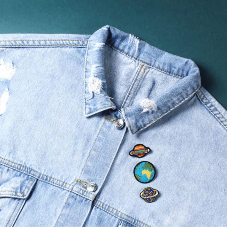 May include: A light blue denim jacket with distressed collar details and silver-toned metal buttons. Three embroidered patches are attached to the front, featuring a colorful planet with rings, a circular Earth globe, and a small dark blue planet.
