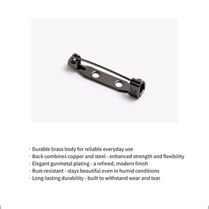 May include: A gunmetal-colored metal brooch pin featuring a long, thin needle mechanism attached to a base with two circular holes. The pin has a dark, reflective finish and a small, rectangular hinge component on one end with a letter M engraved.
