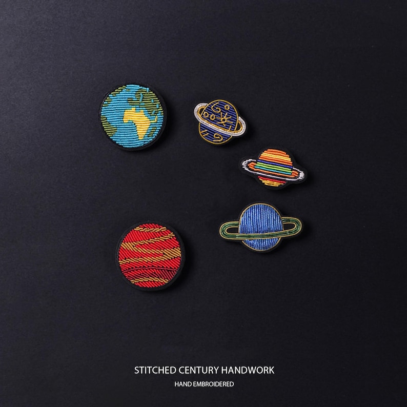 May include: Five circular and oval embroidered patches featuring celestial bodies including a blue and green Earth, a red and yellow striped planet, and three ringed planets in blue, rainbow, and dark blue. Text reads STITCHED CENTURY HANDWORK.