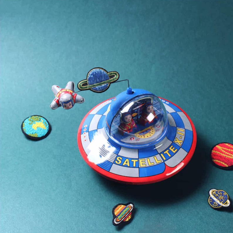 May include: A blue and red tin toy flying saucer labeled SATELLITE X-II with a clear dome containing two figures. A small astronaut figure on a wire orbits the saucer, accompanied by several embroidered planet and space-themed patches.
