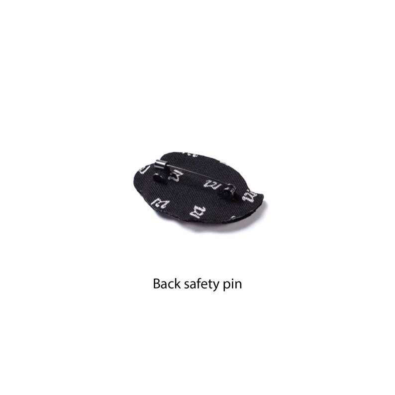 May include: A black fabric brooch with a repeating white letter M pattern. The back of the brooch features a metal safety pin mechanism attached to the textile surface, allowing it to be fastened to clothing or accessories as a decorative item.