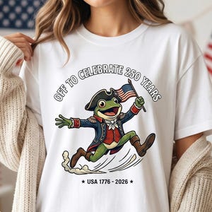 Puede incluir: Camiseta blanca con una rana de dibujos animados vestida con ropa colonial, sosteniendo una bandera estadounidense. La camiseta tiene el texto "OFF TO CELEBRATE 250 YEARS" y "USA 1776-2026". La rana está corriendo, con una estela de humo detrás.