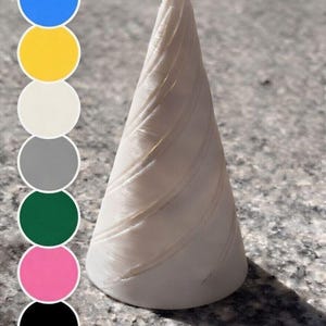 Könnte beinhalten: Ein weißes, kegelförmiges Objekt mit spiralförmigen Rillen, das einem Einhornhorn ähnelt. Das Horn befindet sich auf einer gesprenkelten grauen Oberfläche, mit einer Farbpalette aus blauen, gelben, weißen, grauen, grünen, rosa und schwarzen Kreisen im Hintergrund.