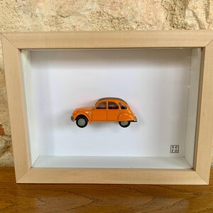 Könnte beinhalten: Ein orangefarbenes Spielzeugauto in einem Schattenboxrahmen. Das Auto hat ein schwarzes Dach und schwarze Reifen. Der Rahmen besteht aus hellem Holz und hat einen weißen Hintergrund. Das Auto ist mittig im Rahmen platziert.