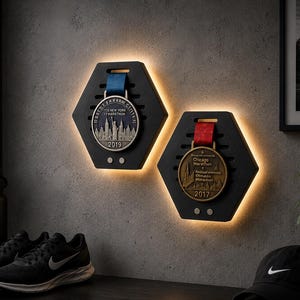 Pode incluir: Dois expositores hexagonais pretos retroiluminados para medalhas de maratona. Um tem uma medalha de 2019, o outro de 2017. Um par de sapatos de corrida pretos e um boné Nike são visíveis.