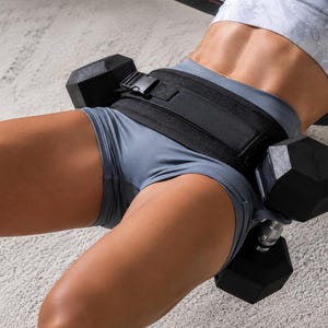 Puede incluir: Un cintur&oacute;n de pesas negro con hebilla est&aacute; asegurado alrededor de la cintura. Dos mancuernas negras est&aacute;n unidas al cintur&oacute;n. La persona lleva pantalones cortos grises y un sujetador deportivo blanco. El fondo es una alfombra gris claro.