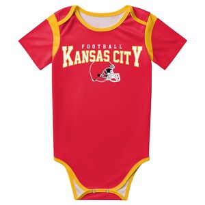 May include: A red short-sleeve baby bodysuit with yellow trim on the neckline, sleeves, and leg openings. The chest features the text "KANSAS CITY" in yellow block letters above a white football helmet graphic with the word "FOOTBALL" above it.