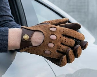 Peut inclure: Gants de conduite en daim beige avec des perforations sur les doigts et les articulations, une ouverture sur le dos de la main et une bride de poignet en cuir marron foncé fixée par un bouton-pression métallique doré circulaire.