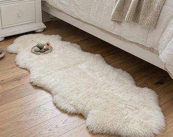 Peut inclure: Un tapis en peau de mouton blanc cassé de forme allongée avec une texture épaisse et duveteuse. Le tapis présente une forme naturelle irrégulière et est posé sur un parquet en bois clair, le long d'un cadre de lit blanc et d'une table de chevet.