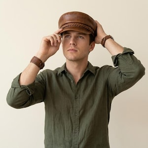 A brown leather baker boy cap featuring a structured crown, a short stiff peak, and a decorative braided leather band across the front. The cap has a smooth finish and a classic rounded shape, providing a clean and uniform appearance.