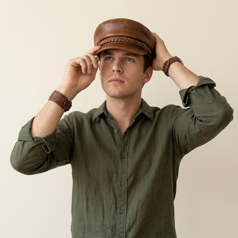 A brown leather baker boy cap featuring a structured crown, a short stiff peak, and a decorative braided leather band across the front. The cap has a smooth finish and a classic rounded shape, providing a clean and uniform appearance.