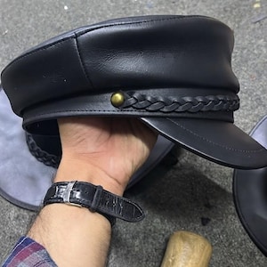 A black leather military-style cap featuring a structured crown, a stiff visor, and a decorative braided leather band across the front. The band is secured on each side by a small, round, metallic brass-coloured stud detail.