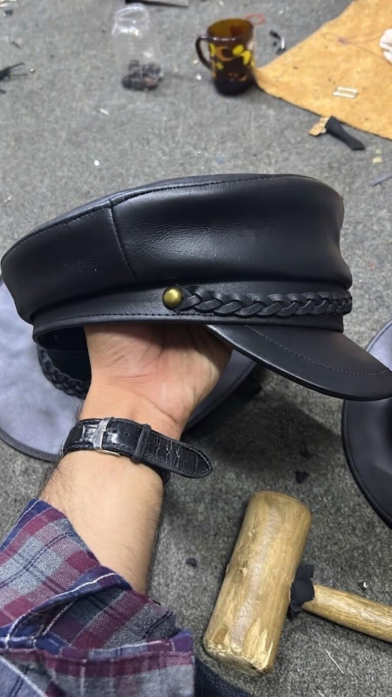 A black leather military-style cap featuring a structured crown, a stiff visor, and a decorative braided leather band across the front. The band is secured on each side by a small, round, metallic brass-coloured stud detail.