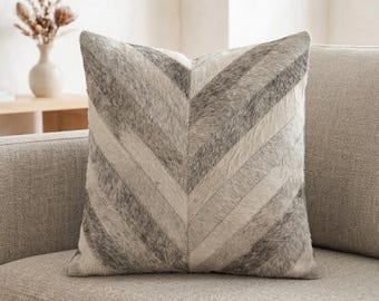 Peut inclure: Un coussin carré avec un motif à chevrons composé de bandes alternées de poils texturés gris clair et gris foncé. Le coussin présente une couture verticale centrale où les rayures diagonales se rejoignent pour former un motif en V.