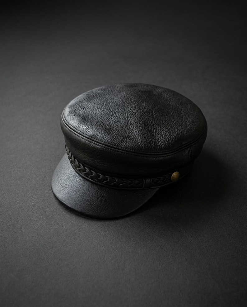 A black leather cap with a textured surface, featuring a braided leather band across the front and a small metallic button detail on the side. The cap has a rounded crown and a short, stiff brim, showcasing a consistent dark finish.
