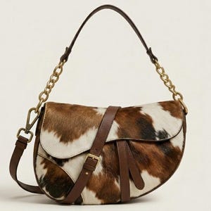 May include: A saddle-shaped shoulder bag featuring a brown and white cowhide pattern with smooth brown leather trim. The bag includes a gold-tone metal chain strap, a matching brown leather shoulder strap, and a central leather buckle strap detail.