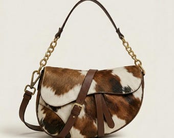 Peut inclure: Un sac à bandoulière en forme de selle avec un motif peau de vache marron et blanc et des garnitures en cuir marron lisse. Le sac comprend une chaîne en métal doré, une bandoulière en cuir marron assortie et une sangle à boucle centrale.