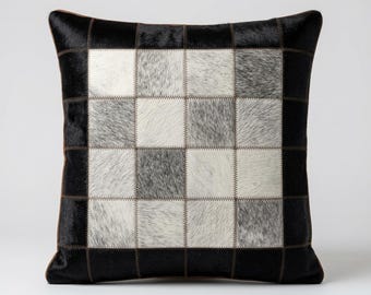 Peut inclure: Un coussin carré composé d'un assemblage de carrés en cuir poilu noir, blanc et gris. Le centre présente un motif en damier blanc et gris, entouré d'une bordure noire unie, le tout assemblé par des coutures apparentes sur chaque pièce.