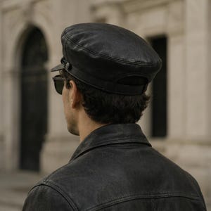 A black leather baker boy cap featuring a structured crown, a short stiff brim, and a decorative black cord detail secured by small buttons on each side. The cap is crafted from smooth black leather with visible stitching.