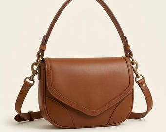 Peut inclure: Un sac à bandoulière en forme de selle en cuir brun clair, doté d'un rabat incurvé avec des détails surpiqués, d'une poignée supérieure unique et de ferrures en métal couleur laiton antique, incluant des clips latéraux et des attaches.