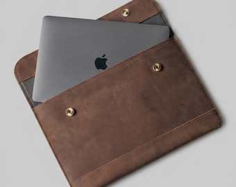 Peut inclure: Une housse d'ordinateur portable en cuir marron avec un ordinateur portable gris à l'intérieur. La housse a une forme rectangulaire avec des coins arrondis et est fixée par deux fermetures à pression couleur laiton. L'ordinateur portable a un logo Apple noir.