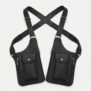 May include: A black dual-pouch shoulder holster bag featuring adjustable elastic straps that cross at the back. Each pouch includes a vertical zippered compartment and a smaller front pocket with a snap button closure, accented with metallic hardware.