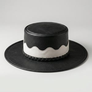 May include: A black leather hat with a flat crown and a wide brim. The base of the crown features a white scalloped leather band layered over the black leather, accented by a braided black leather trim that encircles the bottom of the crown.