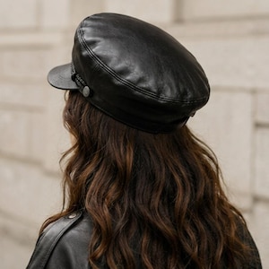 A black leather baker boy cap featuring a structured crown, a short stiff brim, and a decorative braided cord trim across the front. The cap is accented with small circular metal buttons on each side where the cord attaches.