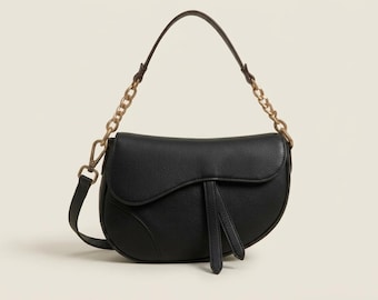 Peut inclure: Un sac à bandoulière noir en forme de selle avec une finition texturée, doté d'un rabat incurvé, de deux languettes en cuir pendantes et d'une anse combinant une sangle noire unie et une section de chaîne en métal doré de chaque côté.