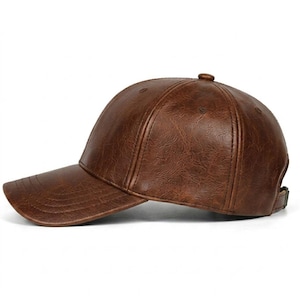 A brown baseball cap with a textured, faux-leather finish. The cap features a six-panel construction with a curved brim, a button at the top, and an adjustable strap with a metal buckle at the back for a secure and comfortable fit.