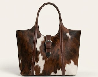 Peut inclure: Un sac cabas avec un motif peau de vache marron et blanc, des garnitures en cuir marron foncé, deux lanières verticales en cuir et une sangle de fermeture centrale avec une boucle en métal argenté et une pointe métallique à l'extrémité.