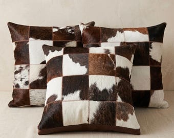 Peut inclure: Trois coussins carrés présentant un motif en patchwork composé de carrés de peau de vache marron, blanc et noir. Chaque coussin est fabriqué à partir de pièces de peau individuelles cousues ensemble avec un fil visible, formant un damier.