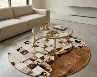 Peut inclure: Un tapis rond composé d'un assemblage de carrés en peau de vache dans des tons blancs, bruns et noirs. Le tapis présente un mélange de cuir brun uni et de motifs en peau tachetée, disposé sous une table basse circulaire en verre.
