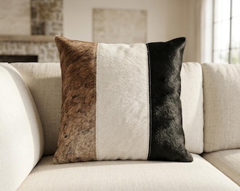 Peut inclure: Un coussin carré présentant un design tricolore avec des panneaux verticaux en peau animale texturée marron, un panneau central en peau blanche et un panneau en peau noire unie sur le côté droit, assemblés par des coutures visibles.